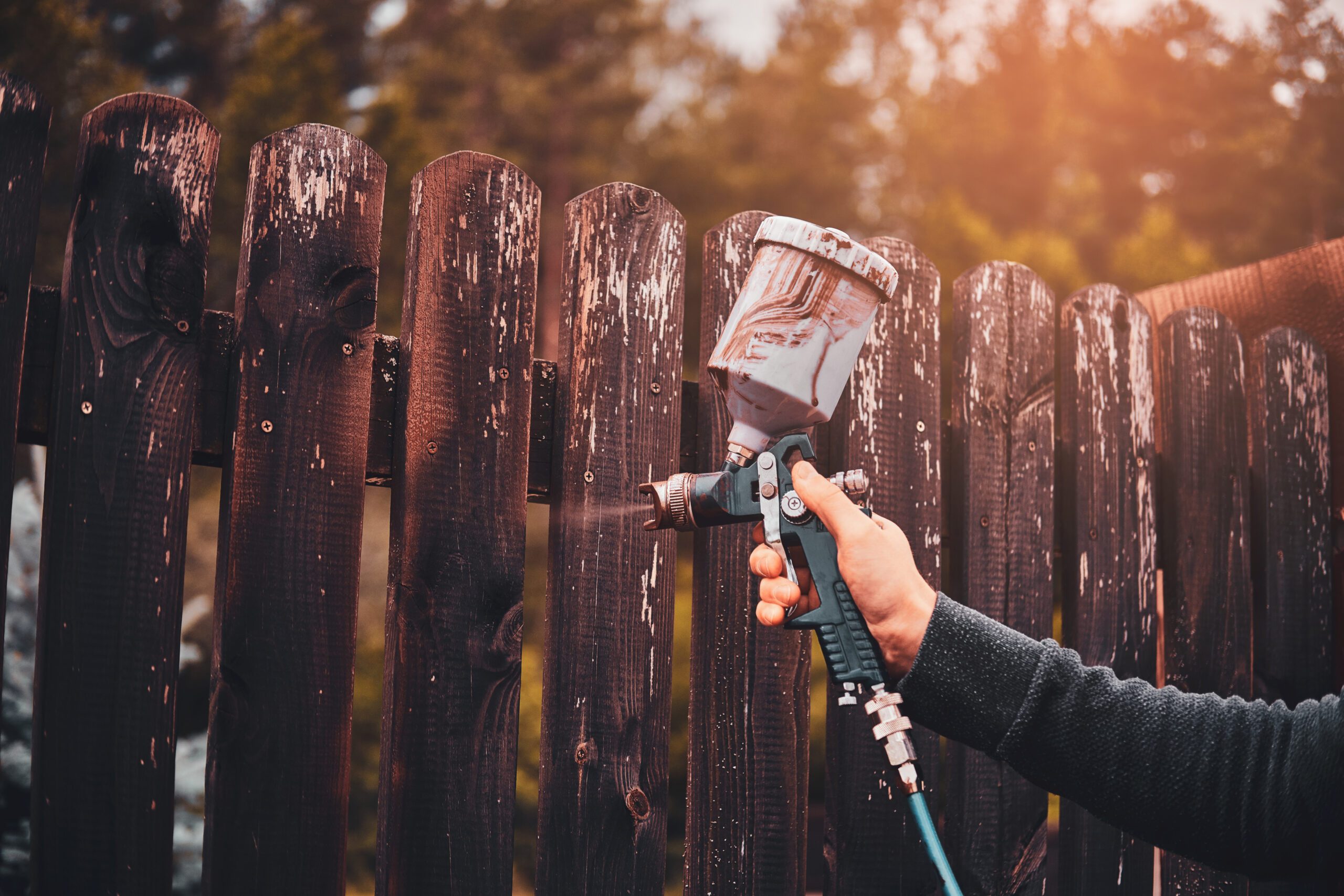 Man is applying paint to the old fence using airbrush at his garden.